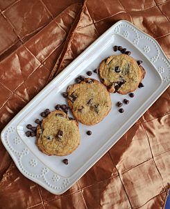 A white platter with three cookies on it and chocolate chips sprinkled on the platter.