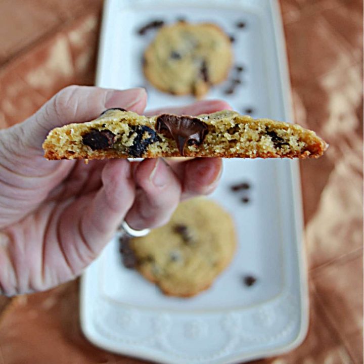 A close up view of a chocolate chip cookie with a bit out of it and a blurred image of a platter of cookies behind it.