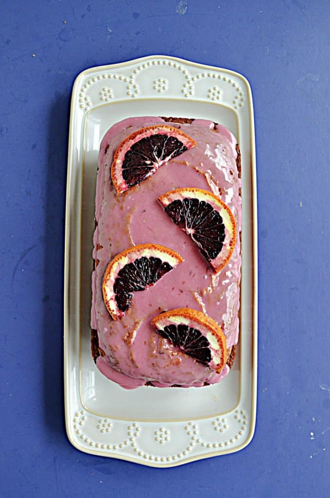 A white platter with a Blood Orange Cake with pink icing and fresh blood oranges on top.