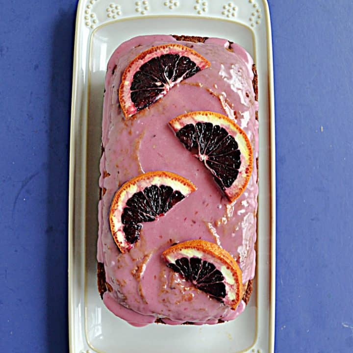 A white platter with a Blood Orange Cake with pink icing and fresh blood oranges on top.