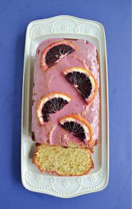 A white platter with a Blood Orange Cake with pink icing and fresh blood oranges on top with a slice of the cake showing.