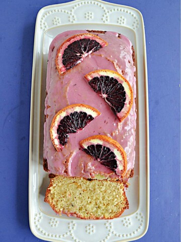 A white platter with a Blood Orange Cake with pink icing and fresh blood oranges on top with a slice of the cake showing.