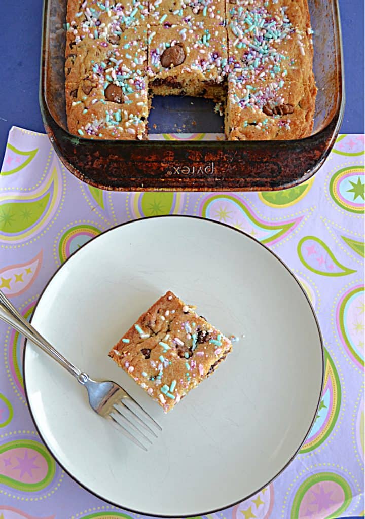 A plate with a Cadbury Creme Egg Blondie square on it along with a fork with a pan of blondies sitting behind it with a piece missing. 