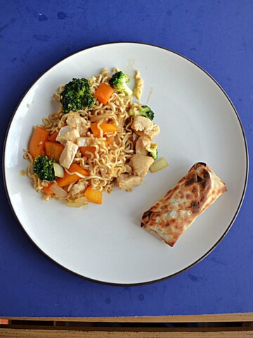 A plate with a large serving of chicken, vegetable, and ramen noodle stir fry with an egg roll on the side.