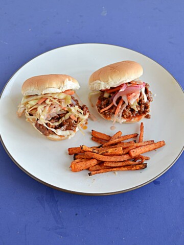 A plate with two Korean BBQ Sloppy Joes topped with pickled cabbage and carrots and a side of sweet potato fries.