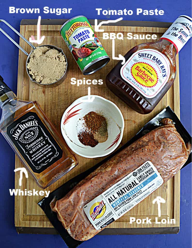 A cutting board topped with a can of tomato paste, a bottle of BBQ sauce, a cup of brown sugar, a bowl of spices, a pork loin, and a bottle of whiskey.