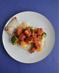 A plate topped with rice and sweet and spicy Firecracker Tofu with peppers and broccoli and an egg roll on the side.