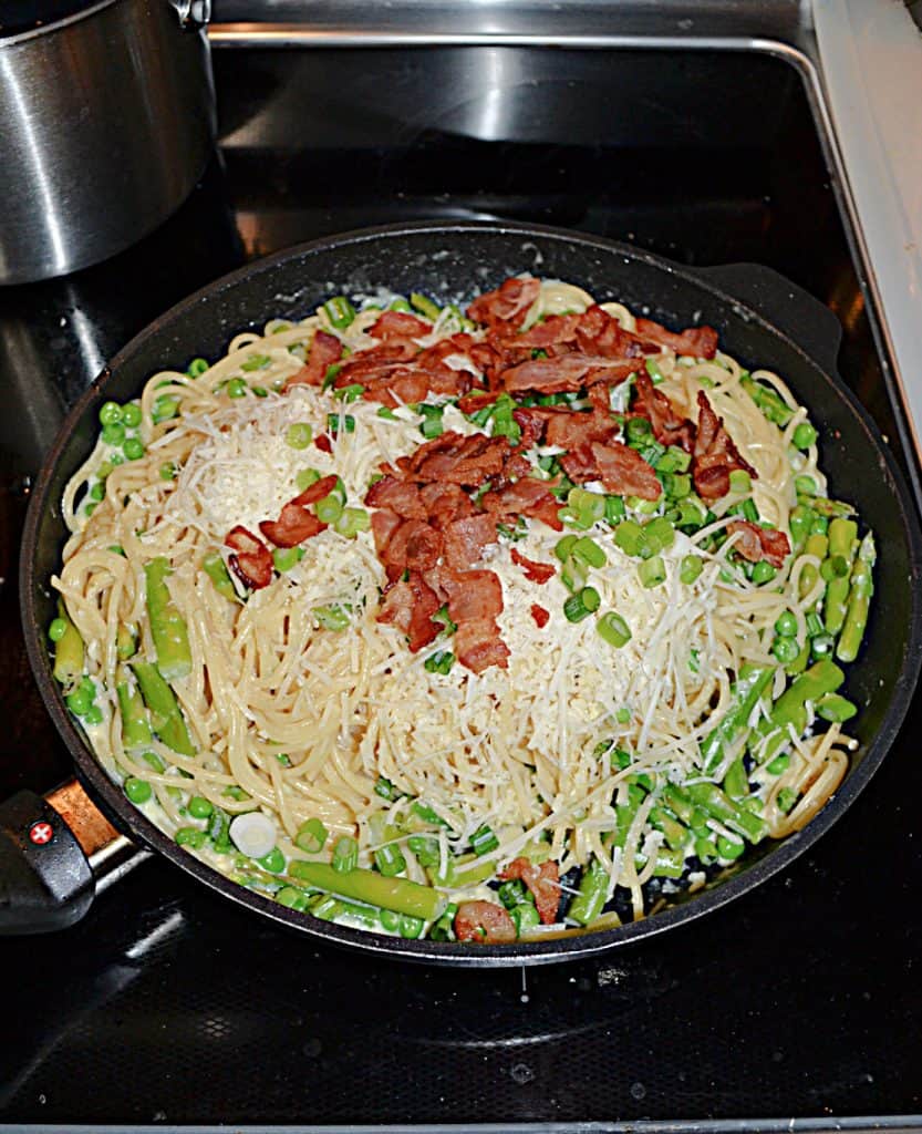A skillet piled high with spaghetti, asparagus, peas, and bacon on top along with parmesan cheese.