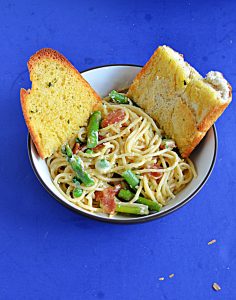 A bowl of spaghetti mixed with with bacon, asparagus, cheese, and peas with two pieces of garlic bread in the bowl.