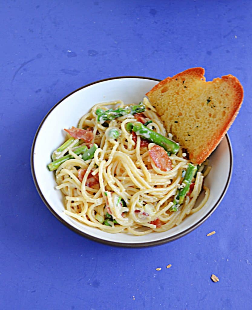 A bowl of spaghetti mixed with with bacon, asparagus, cheese, and peas with one piece of garlic bread in the bowl.
