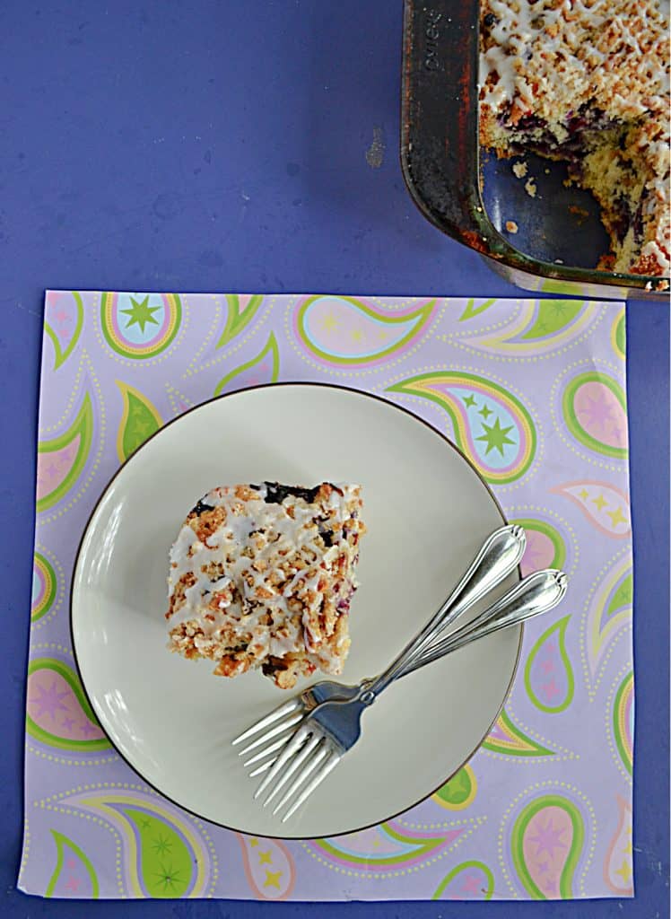 A plate with a slice of blueberry buckle on it and two forks with a corner of the cake pan showing.