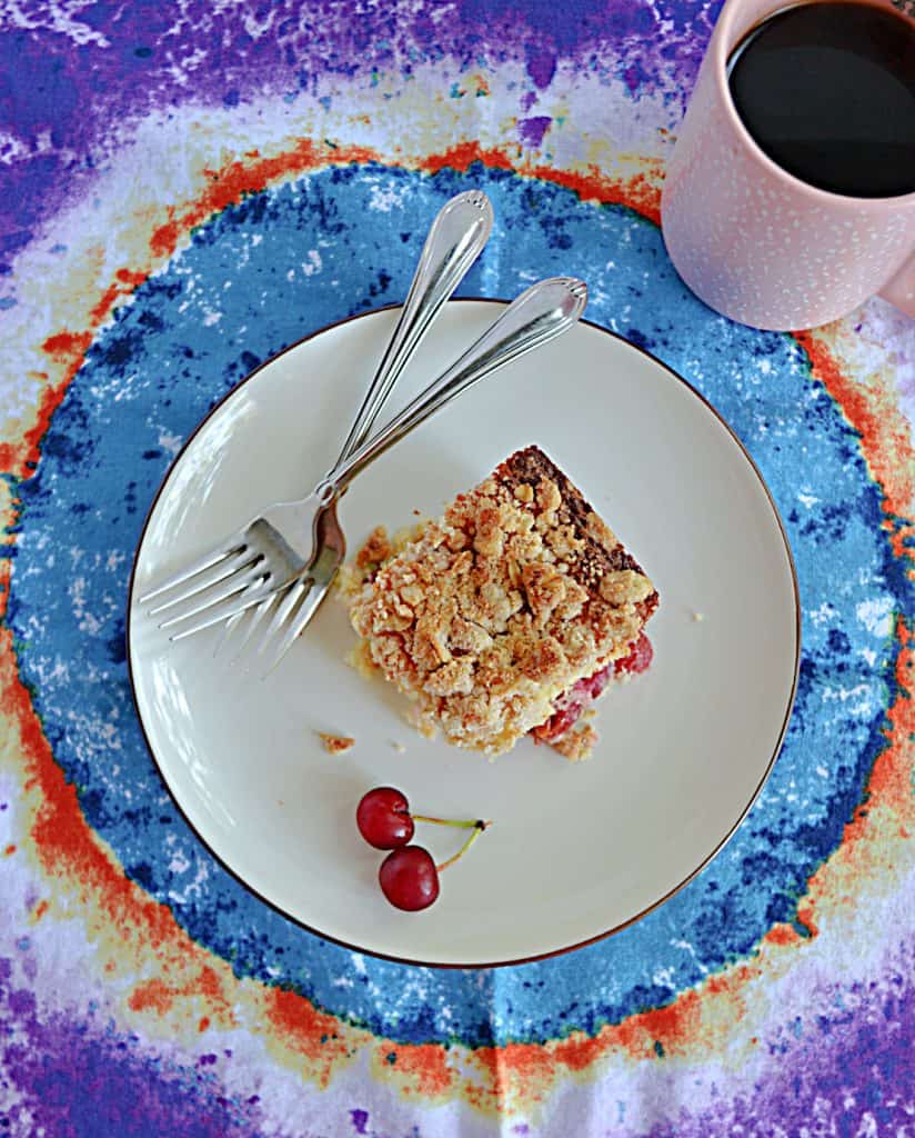 A plate with a piece of sour cherry coffee cake, a pair of sour cherries, 2 forks, and a cup of coffee on the side.