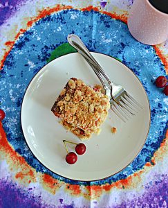 A top view of A plate with a piece of sour cherry coffee cake, a pair of sour cherries, 2 forks, and a cup of coffee on the side.