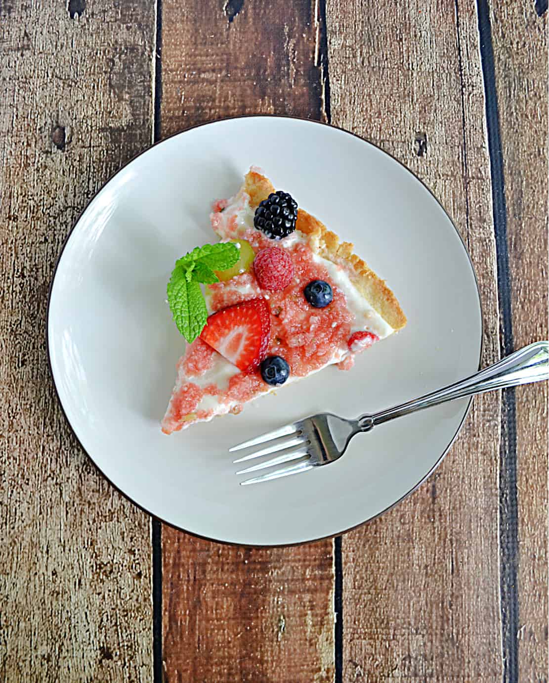 A plate with a slice of fruit pizza topped with strawberries, blueberries, and a mint left with a fork on the plate.