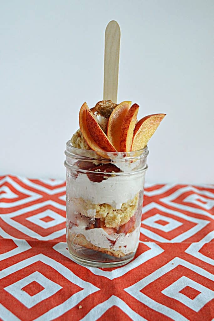 A jar layered with cinnamon whipped cream, fresh peaches, and sweet biscuits with peaches on top and a spoon sticking out of the jar.
