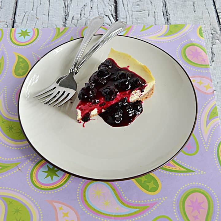 A slice of Vanilla Bean Cheesecake topped with blueberry compote with two forks on the plate.