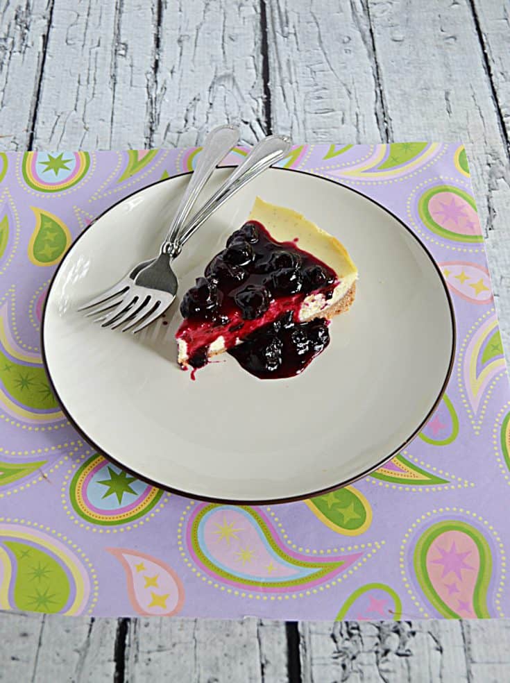 A slice of Vanilla Bean Cheesecake topped with blueberry compote with two forks on the plate.