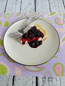 A plate with a slice of Vanilla Bean Cheesecake topped with blueberry compote with two forks on the plate.