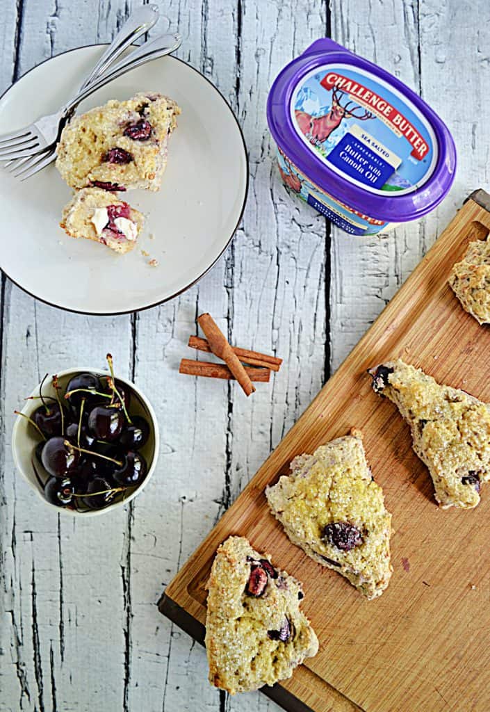 A cutting board with scones on it, a tub of butter, 3 cinnamon sticks, a bowl of cherries, and a plate with a scone and a fork on it. 