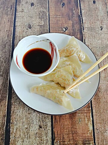 A plate with 4 chicken gyoza dumplings, a pair of chopsticks, and a bowl of dipping sauce on it.