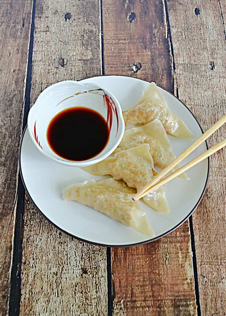 A plate with 4 chicken gyoza dumplings, a pair of chopsticks, and a bowl of dipping sauce on it. 