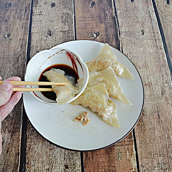A pair of chopsticks dipping a chicken gyoza into a bowl of sauce with three other chicken gyoza dumplings on a plate next to it.