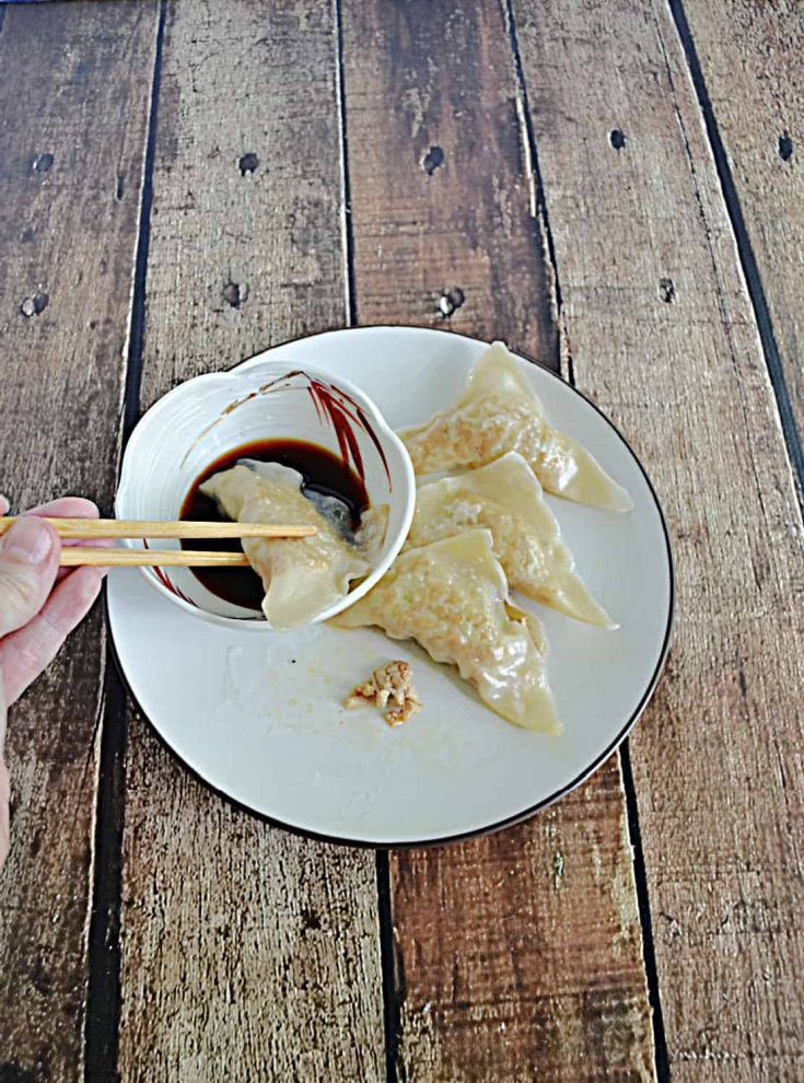 A pair of chopsticks dipping a chicken gyoza into a bowl of sauce with three other chicken gyoza dumplings on a plate next to it.