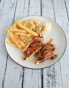 A plate with crinkle fries, cauliflower, and two barbecue chicken tenders topped with bacon onion jam.