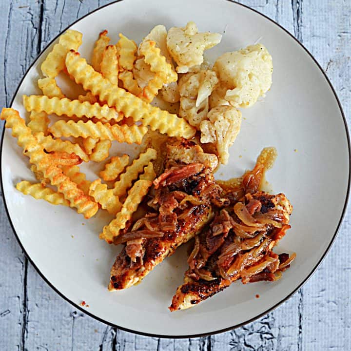 A close up view of A plate with crinkle fries, cauliflower, and two barbecue chicken tenders topped with bacon onion jam.