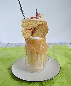 A milkshake with a thick caramel rim topped off with whipped cream, a graaham cracker, a slice of cake, and two straws.