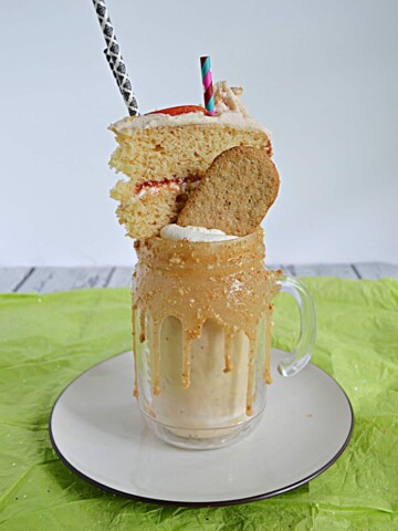 A milkshake with a thick caramel rim topped off with whipped cream, a graaham cracker, a slice of cake, and two straws.
