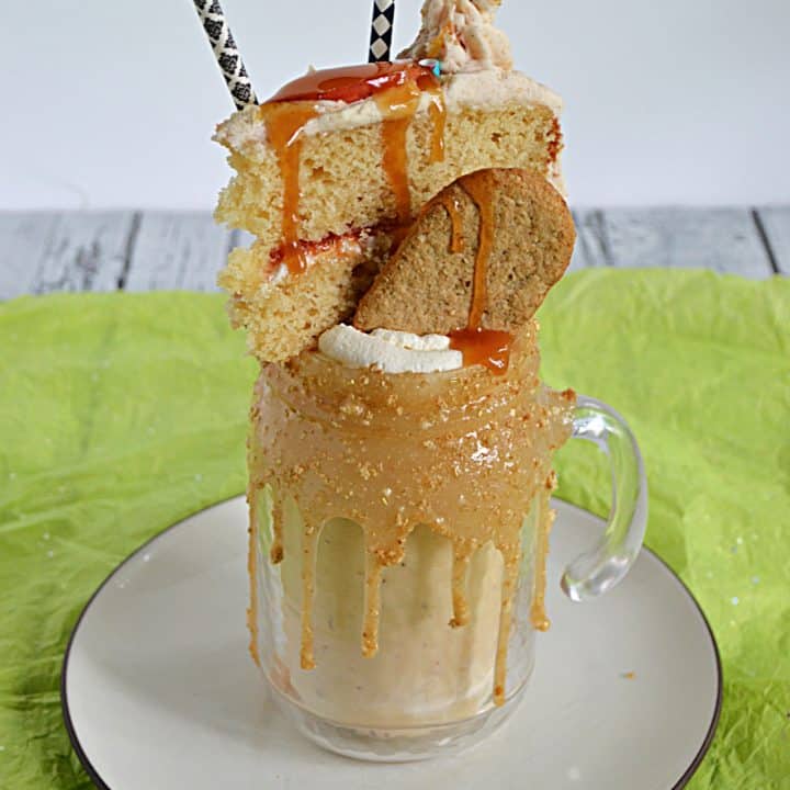 A milkshake sitting in a glass with a thick caramel rim them topped with a graham cracker, a slice of cake, and two straws.