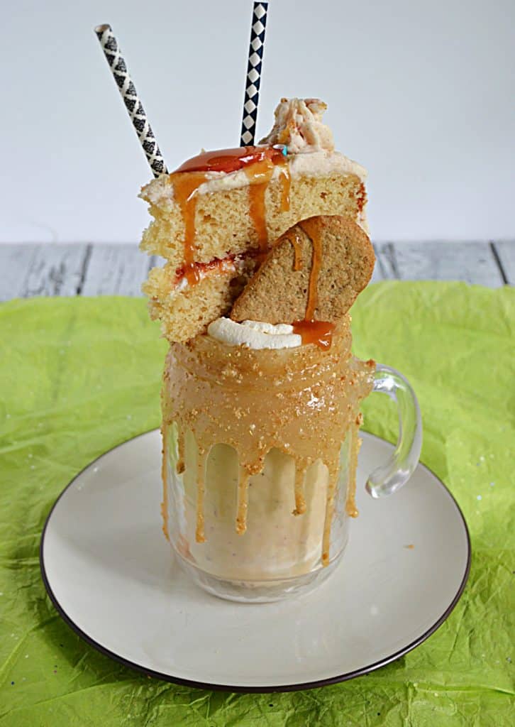 A milkshake sitting in a glass with a thick caramel rim them topped with a graham cracker, a slice of cake, and two straws. 