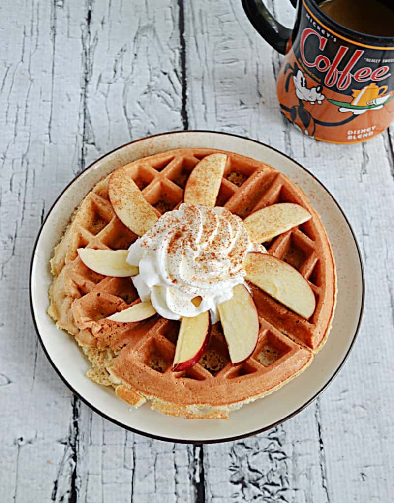 A plate with a large Apple Spice Cake Mix Waffle on it topped with apple slices, whipped cream, and cinnamon on top. 