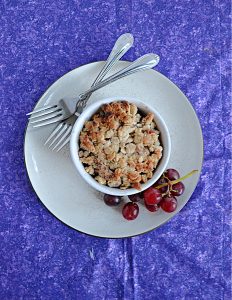 A plate with a ramekin of grape crumble on it, a pile of grapes, and two forks.