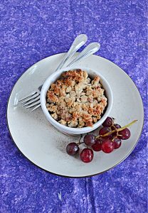 A plate with a bowl of grape crumble on it with a stem of grapes on the one side and two forks on the other side.