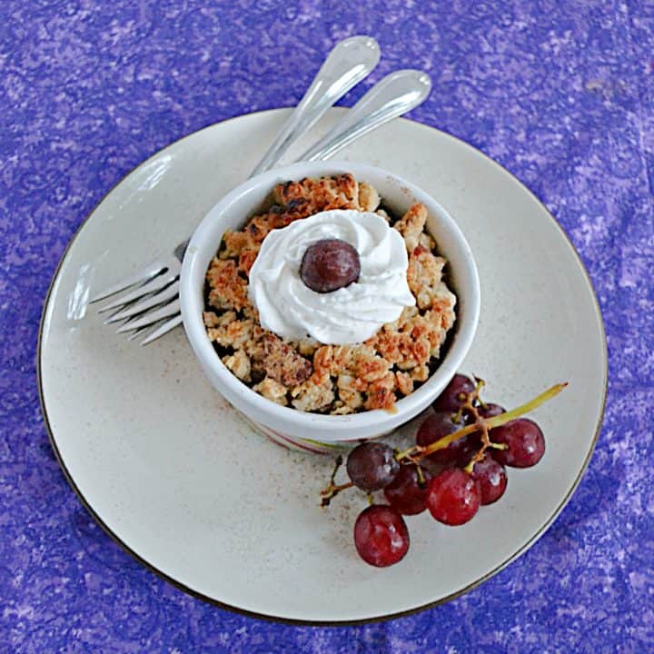 A plate with a bowl of grape crumble on it topped with a grape and whipped cream, a stem of grapes on the one side and two forks on the other side.