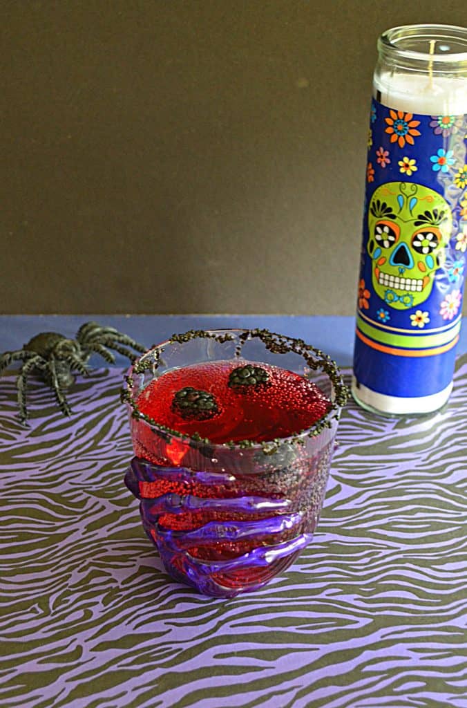 A glass with a skeleton hand on it filled with red Vampire's Kiss Cocktail and floating blackberries with a spider and a skull candle in the background.