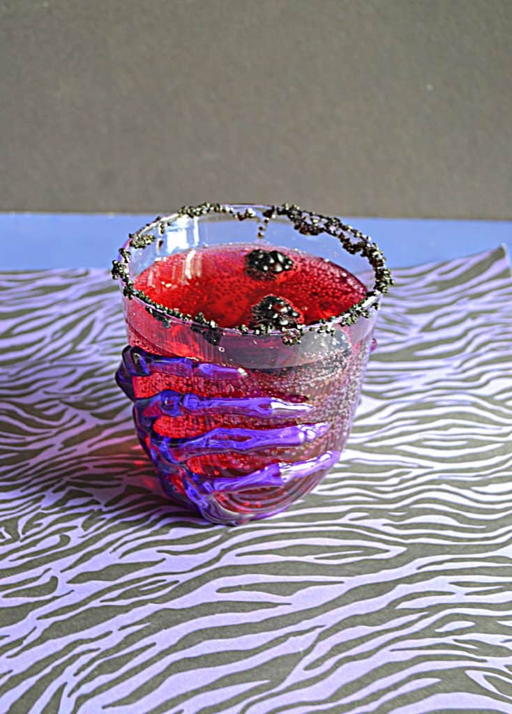 A close up view of a glass with a skeleton hand on it filled with red Vampire's Kiss Cocktail and floating blackberries. 