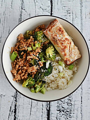 A bowl filled with ground chicken, broccoli, rice, and an egg roll.