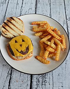 A plate with a piece of cheese that looks like a Jack-o-lantern face on a toasted bun along side a pile of fries.