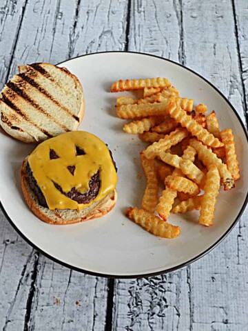 A plate with a piece of cheese that looks like a Jack-o-lantern face on a toasted bun along side a pile of fries.