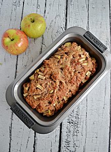 A bread pan with apple banana bread in it with 2 apples placed behind it.