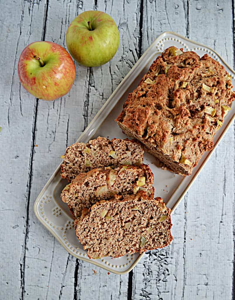 A platter with three slices of apple banana bread and the rest of the loaf with 2 apples in the background.