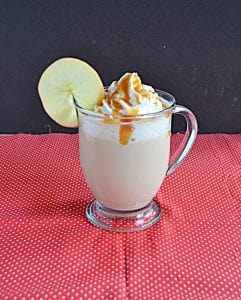 A side view of an Apple Chai Tea Latte with whipped cream and caramel on top and an apple wheel on the lip of the glass.