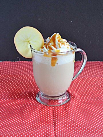 A side view of an Apple Chai Tea Latte with whipped cream and caramel on top and an apple wheel on the lip of the glass.