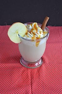 an Apple Chai Tea Latte with whipped cream and caramel on top and an apple wheel on the lip of the glass and a cinnamon stick stuck in it.