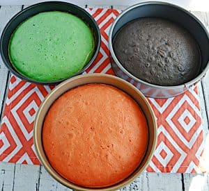 Three cake pans. One with orange cake, one with a green cake, and one with a black cake.