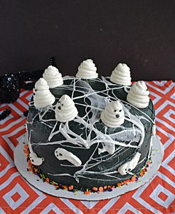 A black cake covered in marshmallow spiderwebs and swirled buttercream ghosts on top.