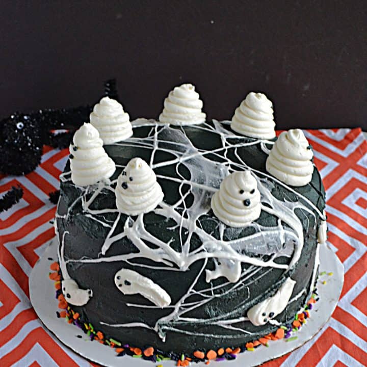 A black cake covered in marshmallow spiderwebs and swirled buttercream ghosts on top.